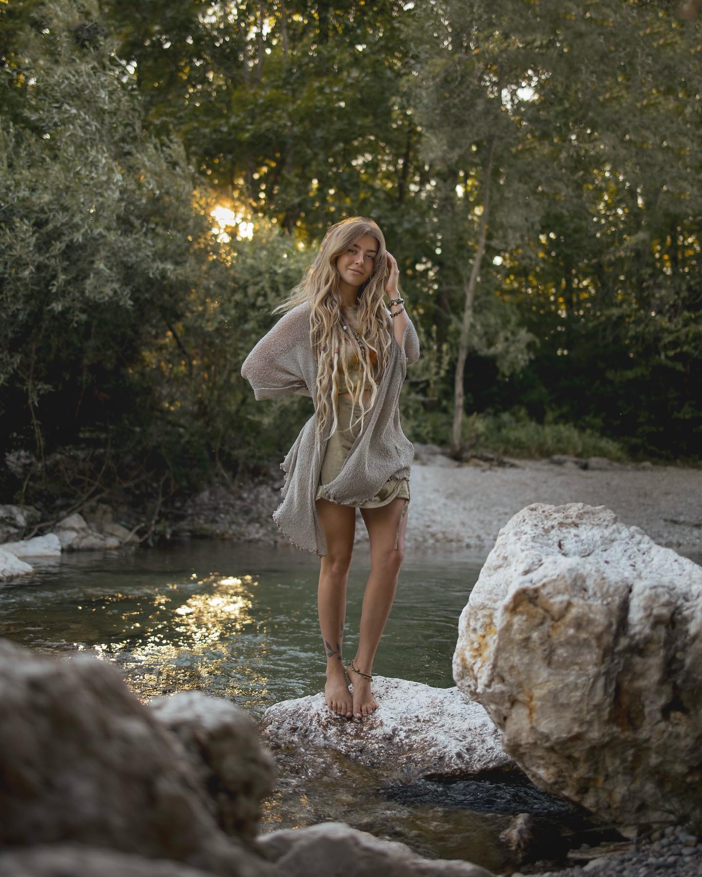 a young woman with long blonde dreadlocks stands barefoot on a rock in a shallow river. she is wearing a light grey cardigan and a short dress. the setting is a tranquil forest with lush green trees and sunlight filtering through the leaves. the overall atmosphere is peaceful and serene. the woman's pose and expression convey a sense of calm and relaxation. the image has a bohemian, freespirited aesthetic.