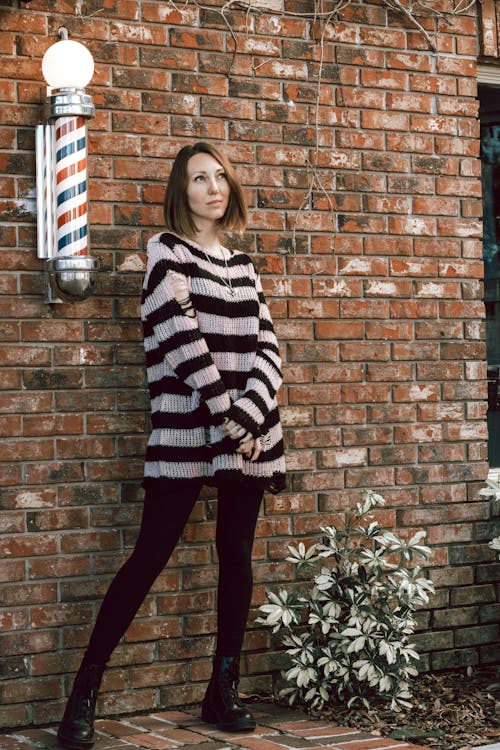 a young woman stands against a brick wall with a barber pole next to her. she is wearing an oversized striped sweater, black leggings, and combat boots. she has shoulderlength brown hair and is looking off into the distance. a small plant is in front of her.