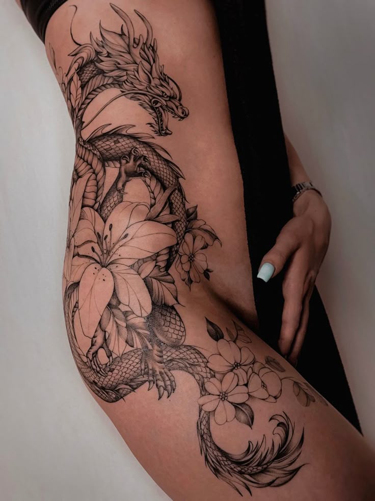 the image shows a tattoo of a dragon intertwined with flowers on a woman's thigh. the tattoo is black and white and features a detailed dragon with scales, an open mouth, and prominent horns. the dragon is surrounded by various flowers, including lilies and smaller blossoms with detailed petals and leaves. the tattoo wraps around the thigh area, extending from the upper thigh down towards the knee. the woman's hand and wrist with a watch are also visible.