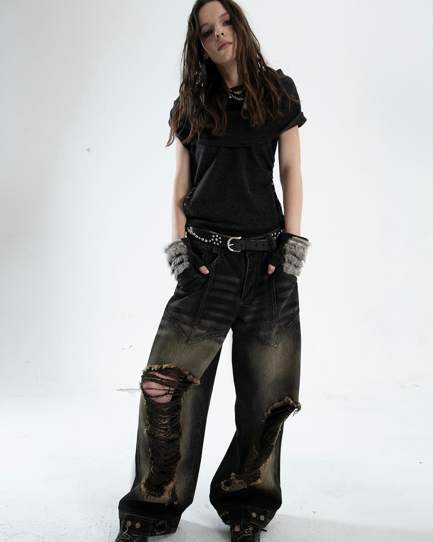 a fullbody shot of a woman model showcasing a fashion outfit against a clean white background. the outfit consists of a dark grey cowlneck shortsleeved top, black wideleg distressed jeans with a black studded belt, and grey fingerless gloves. the model has long dark hair, and she is posed standing with a slightly tilted head and a neutral expression. the focus is on the model's outfit, with the clothing details and styling emphasized. the overall aesthetic leans toward a grunge or alternative style.