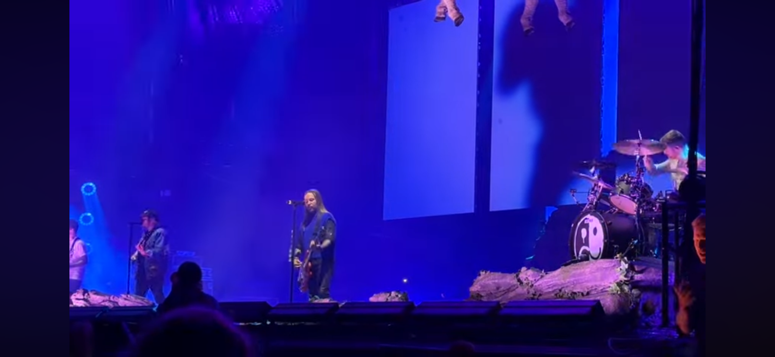 a rock band performs on stage at a concert. the lead singer is in the center, playing a guitar, and the drummer is on the right. the stage is bathed in blue light, and the audience is visible in the background.