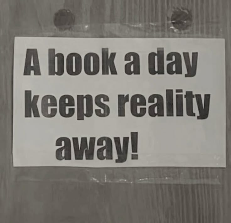 the image shows a sign that reads "a book a day keeps reality away!" in black text on a white background. the sign is attached to a surface with visible staples.