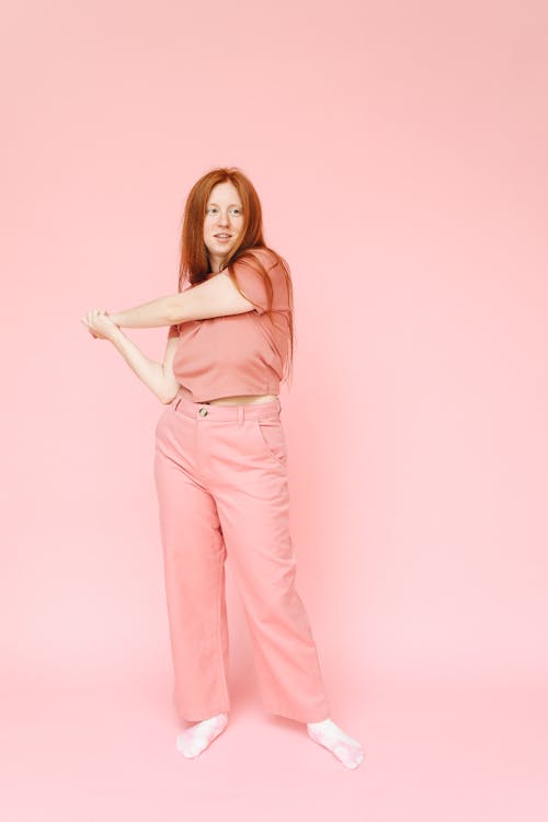 a fulllength studio shot of a young, redhaired caucasian woman standing on a pink background. she's wearing a matching pink crop top and pants, with white socks on her feet. the woman is stretching, her arms crossed in front of her, and she is looking off to the side with a slight smile.