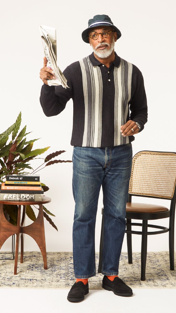 an older african american man with a relaxed and stylish look stands in a studio shot against a white background. he is wearing a black bucket hat, glasses, a black and white striped sweater, jeans, black loafers with orange socks, and is holding a newspaper.  nearby is a chair and small table with books and a plant. the overall impression is one of casual comfort and style.