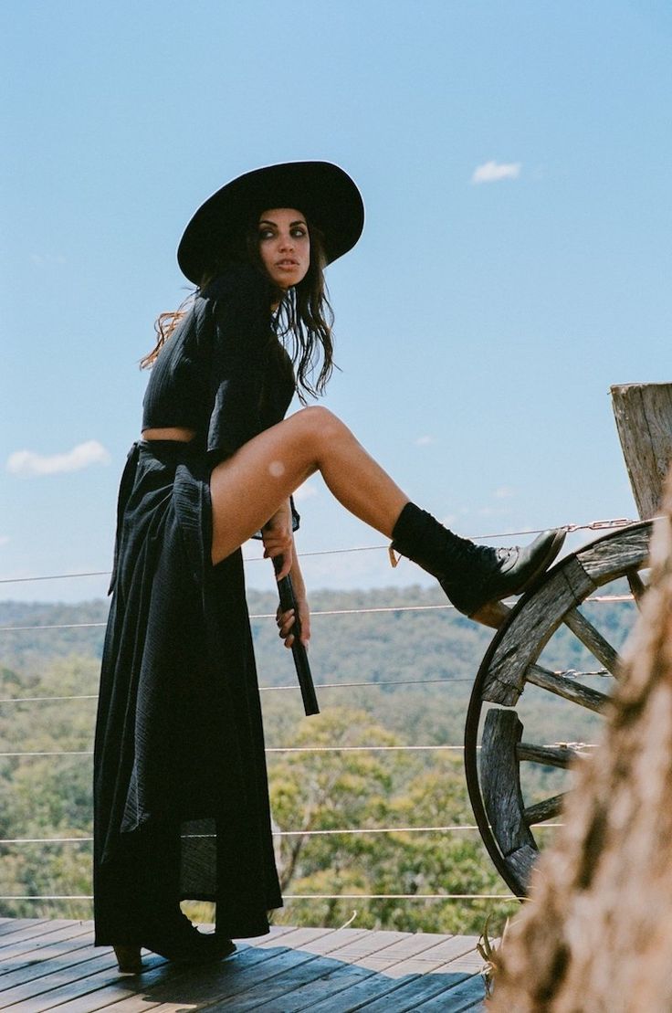 a woman wearing a black dress, hat, and boots is posing outdoors on a wooden railing. she is looking away from the camera, towards the forest and trees in the distance. the sky is blue with some clouds.  the woman is surrounded by nature and is dressed in a stylish and fashionable outfit.