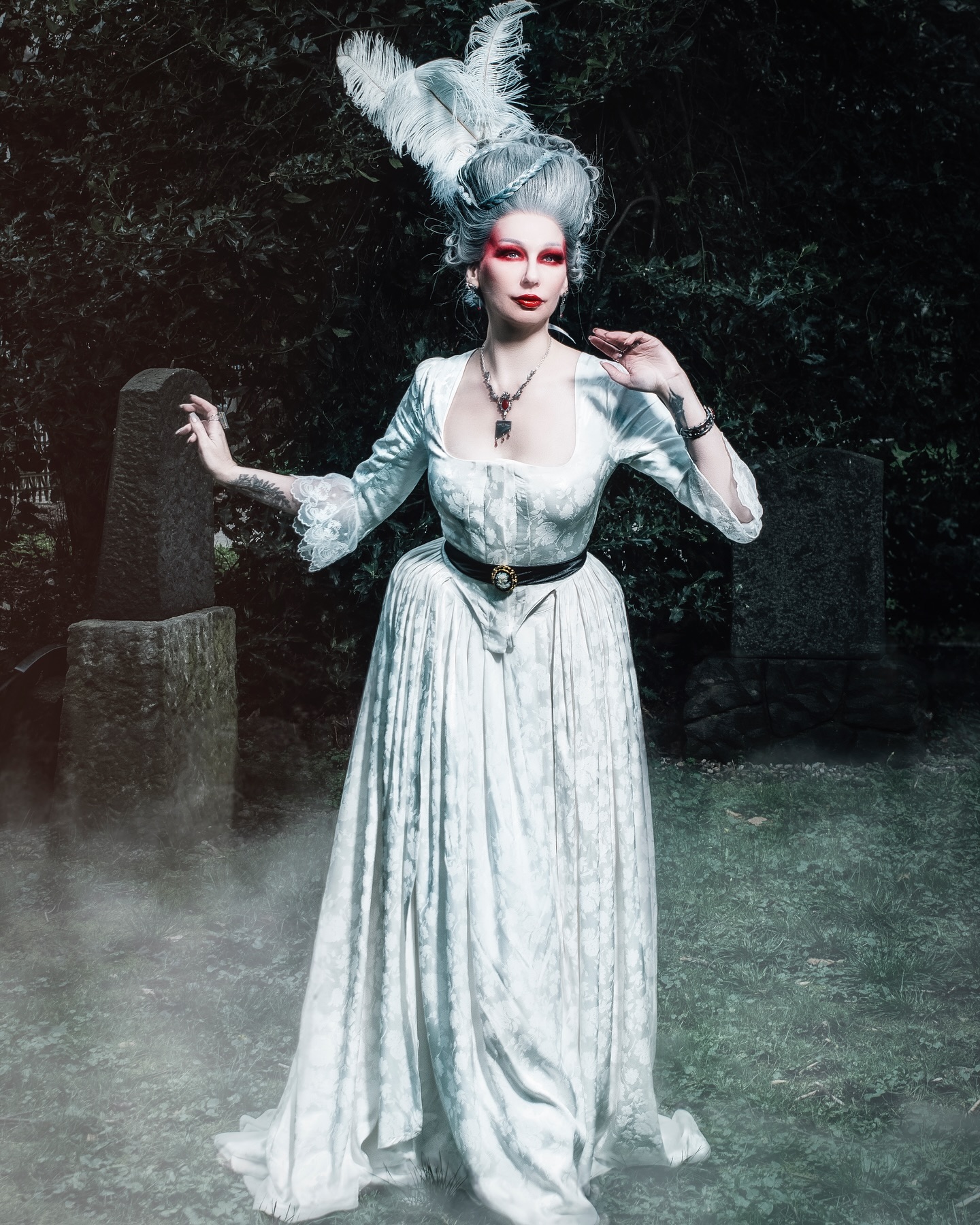 a full shot of a woman in a stunning white rococostyle gown stands in a graveyard. the woman's pale skin and striking red lips contrast sharply with the muted colors of her surroundings. she sports an elaborate white wig adorned with feathers.  the atmosphere is eerie and mysterious, enhanced by the fog swirling around her feet and the somber setting of the ancient tombstones. the image has a dramatic and artistic quality, suggesting a gothic or fantasy theme.