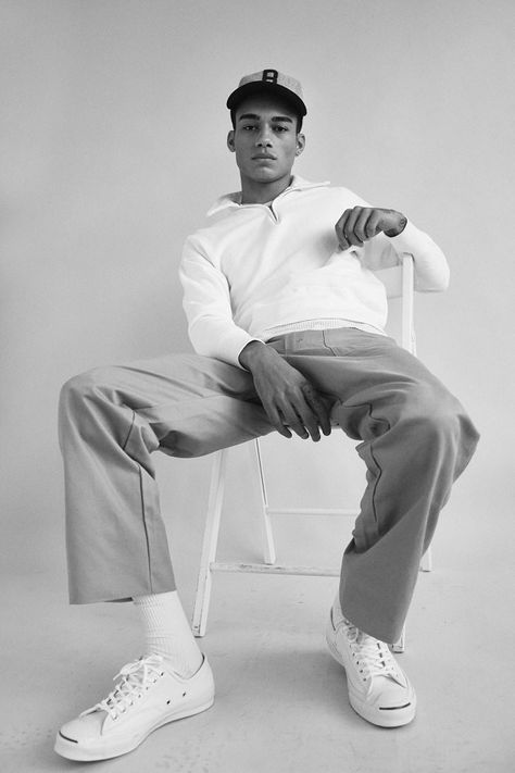 a young man in a white sweater, black cap, and lightcolored pants sits relaxed on a white chair, his white sneakers are visible. the image is taken in a studio and is in black and white.