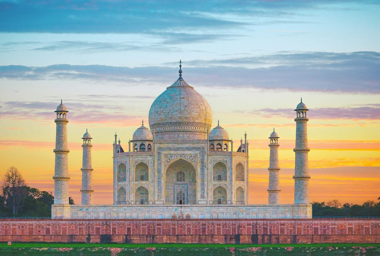 the image shows the taj mahal at sunset. the mausoleum is made of white marble and is located in agra, india. it is a unesco world heritage site. the yamuna river is in the foreground and reflects the building. the sky is filled with orange and pink clouds. there are gardens in front of the taj mahal.