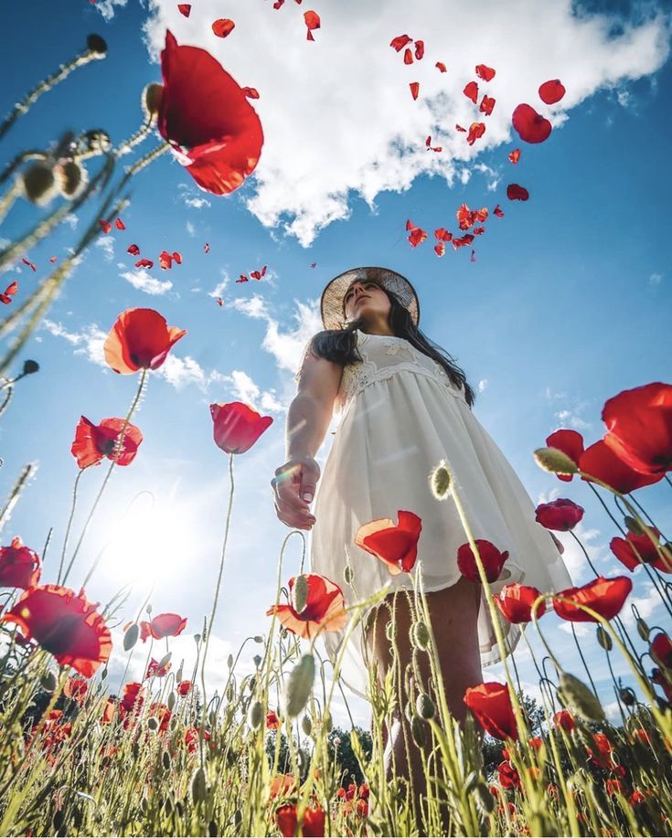 the image shows a young woman in a white dress and straw hat standing in a field of vibrant red poppies.  the photo is taken from a low angle, looking up at her from the ground, with the poppies and sky forming a beautiful backdrop.  petals are falling from the poppies, adding to the ethereal and dreamy quality of the picture. the overall mood is one of summery joy and peaceful serenity. the sun shines brightly, creating a contrast between light and shadow in the field.