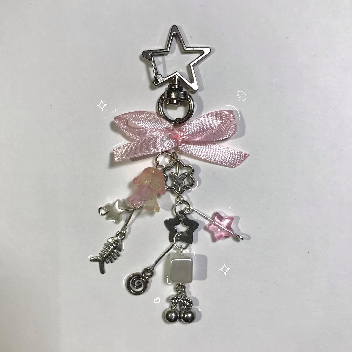 the image shows a keychain with various charms hanging from a pink ribbon tied below a starshaped clip. the charms include a fishbone, a flower, star beads, a square bead, and a pair of cherries.