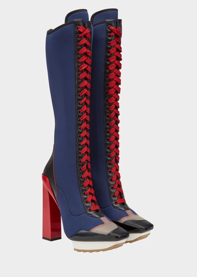a pair of women's highheeled kneehigh boots are shown. the boots are navy blue neoprene with red laces that go up the entire length of the boot. the heel is a red, thick block heel. the toe is a slightly squaredoff shape. the boots are angled slightly to show the side profile. the background is white.