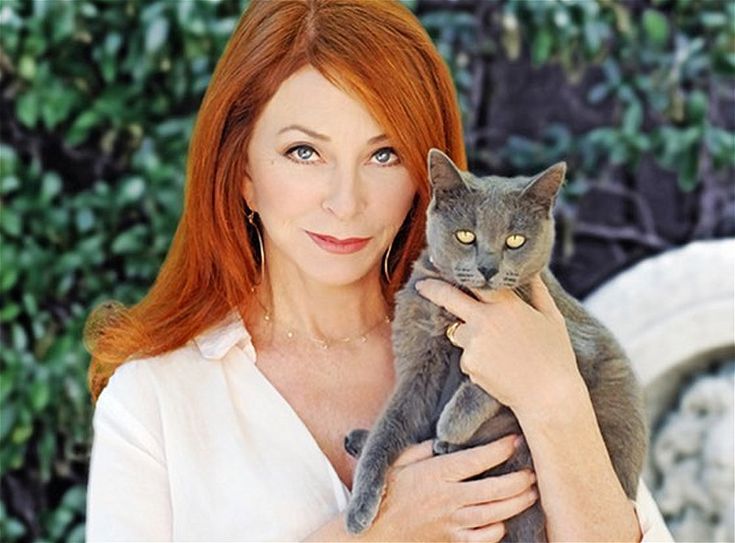 a woman with long, red hair is holding a grey cat close to her. the woman is smiling and looks happy. the cat is resting its head on the woman's shoulder. they are outdoors, in a garden with green leaves. the woman is wearing a white shirt and a necklace.  the cat is a grey cat and has yellow eyes.