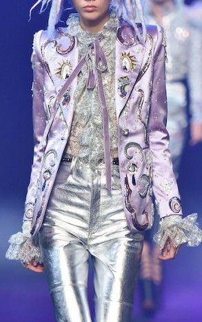a model is walking the runway in a glamorous outfit. she is wearing a silver sequin jacket with purple embroidery. she also has on silver pants and a silver belt. her hair is light and flowing. the background is a purple stage.