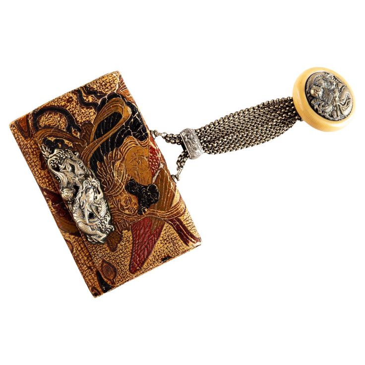 the image shows a vintage, orientalstyle purse with a metallic chain and an ivorycolored clasp. the purse itself is ornate and embroidered with gold and red designs, potentially depicting dragons or other asian motifs. a silver dragon embellishment is attached to the side of the purse. the chain appears to be silver, connected to the purse with a decorative metal piece. the clasp is round, made of ivory, and features a metal detail. the overall style of the purse suggests it may be a vintage or antique fashion accessory.