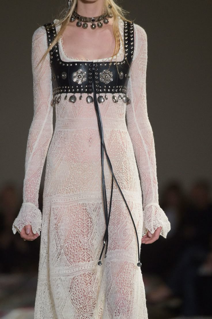 a model is seen wearing a long, creamcolored lace dress with long, slightly ruffled sleeves. over the dress, she wears a short, black leather top adorned with silver studs and floral embellishments. she also has a silver choker around her neck. the dress is semisheer, revealing a hint of skin underneath. the image is likely from a fashion runway event.
