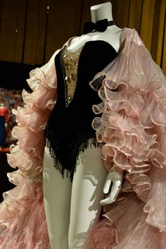a vintage showgirl costume is displayed on a mannequin. the costume features a black velvet corset with intricate beading and a sheer, flowing pink overskirt with layers of ruffles. the bodice of the corset is lowcut and boned, accentuating the feminine form. a black bow tie adorns the mannequin's neck, adding a touch of masculine contrast. the costume is likely from a burlesque or theatrical performance, and it evokes a sense of glamour and opulence. the soft, diffused lighting highlights the delicate textures and intricate details of the garment.