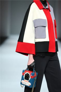 a fashion model walks down the runway wearing a stylish red, white and black coat. she is carrying a small, colorful box bag with a gold chain strap. the model is posing in a stylish way, with her arms down and her legs together. the model is showcasing a modern and chic outfit.