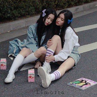 two young asian girls are sitting on a sidewalk together. one girl is wearing a blue jacket and white socks and shoes, and the other is wearing a white shirt and denim shorts with white socks and shoes. they are both smiling and looking at the camera. they are friends and are having fun together.