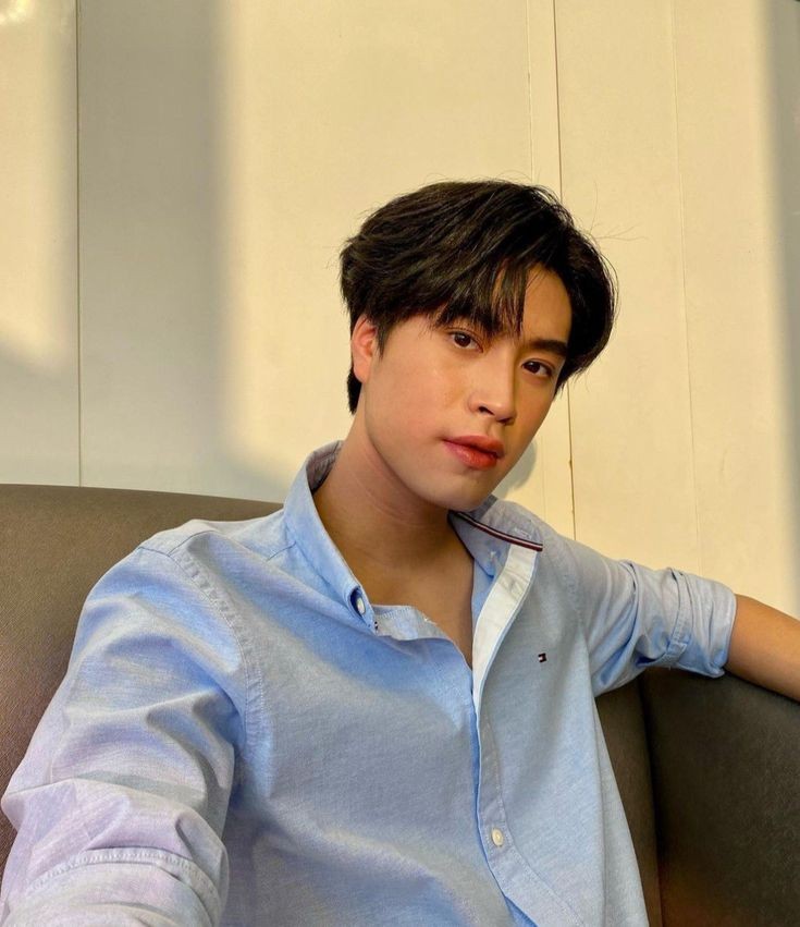 a young asian man with dark hair and a light blue, unbuttoned collared shirt sits on a dark couch, taking a selfie. the background is a simple white wall with vertical lines, and natural light bathes the scene from the left, casting soft shadows. he offers a relaxed, neutral expression towards the camera.