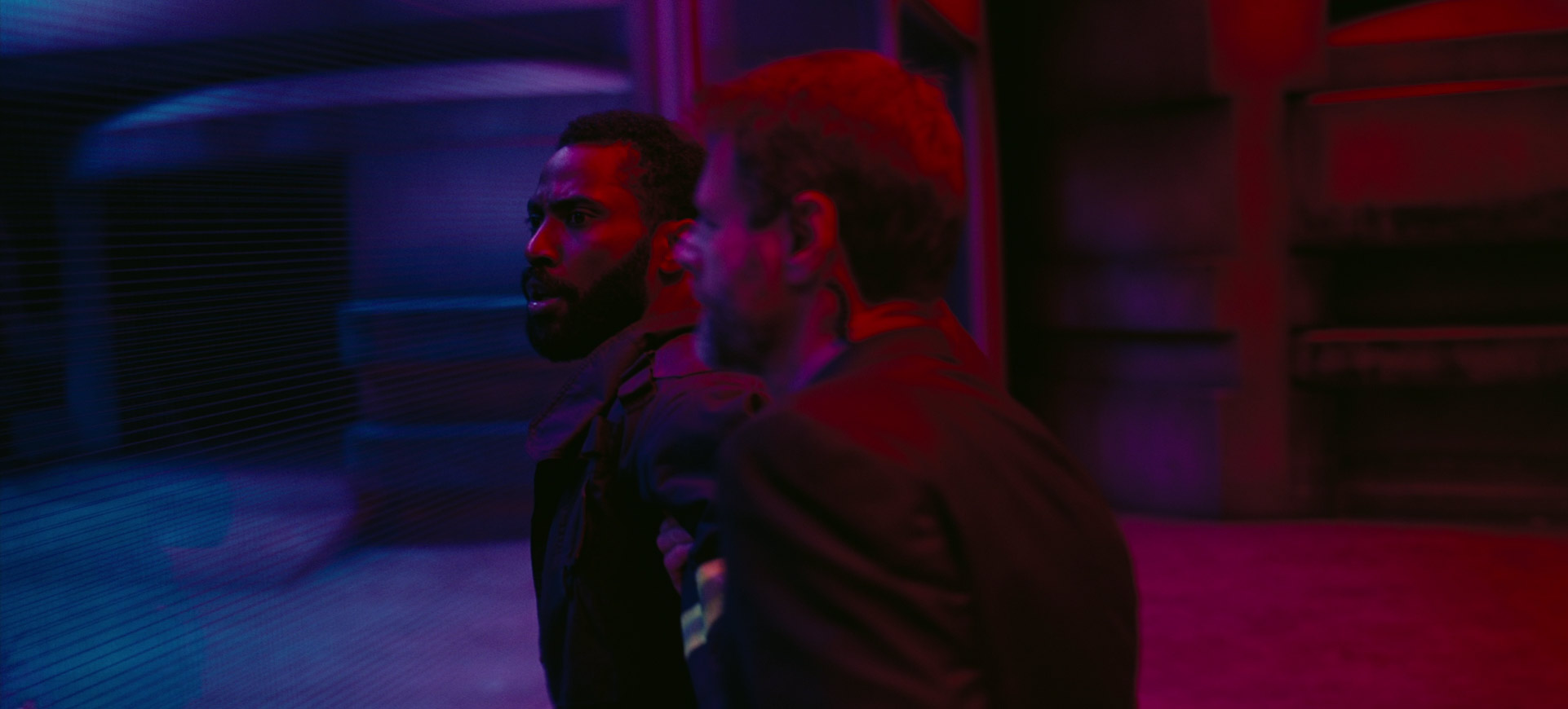 in a dimly lit, futuristic setting, a black man with a beard is being escorted by a person with short brown hair.  the man in the foreground is wearing a dark jacket and looking to the left with a concerned expression.  he appears to be in police custody, although no handcuffs are visible.  the scene is lit with blue and purple neon, creating a tense and dramatic atmosphere.