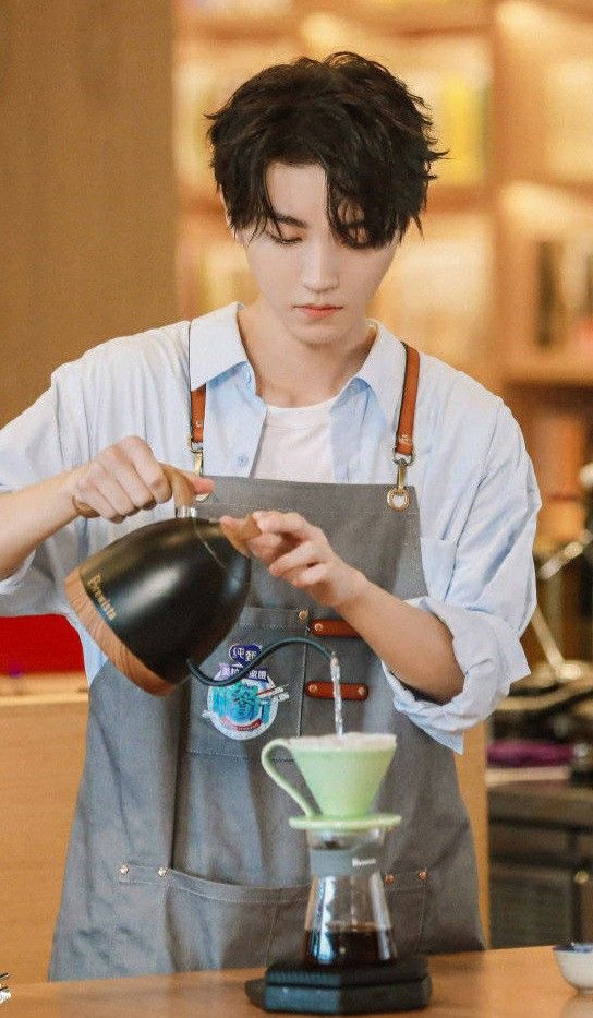 a young man with black hair and wearing a blue shirt and gray apron stands in a coffee shop. he is brightly lit, likely by artificial light.  he is carefully pouring hot water from a black electric kettle over coffee grounds in a green pourover coffee maker. he appears focused on the task, his expression neutral as he prepares the coffee.