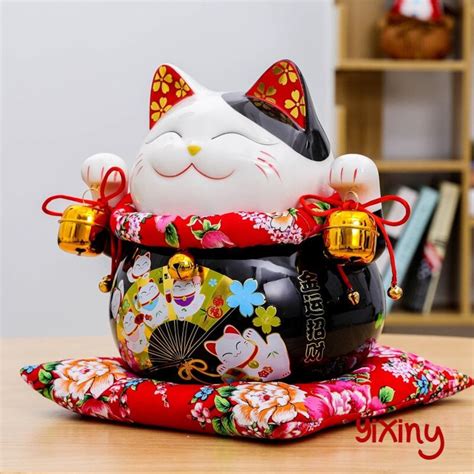 a closeup shot of a maneki neko, or beckoning cat, piggy bank. the cat is predominantly white and black with red floral accents on its ears and a red floral patterned cushion around its neck.  it's sitting on a red floral patterned cushion.  the piggy bank is ornately decorated with gold bells and smaller maneki neko images on its body. the overall style is traditional japanese. the background shows a blurred image of a shelf with books and other objects. the image is welllit and shows the details of the figurine clearly.
