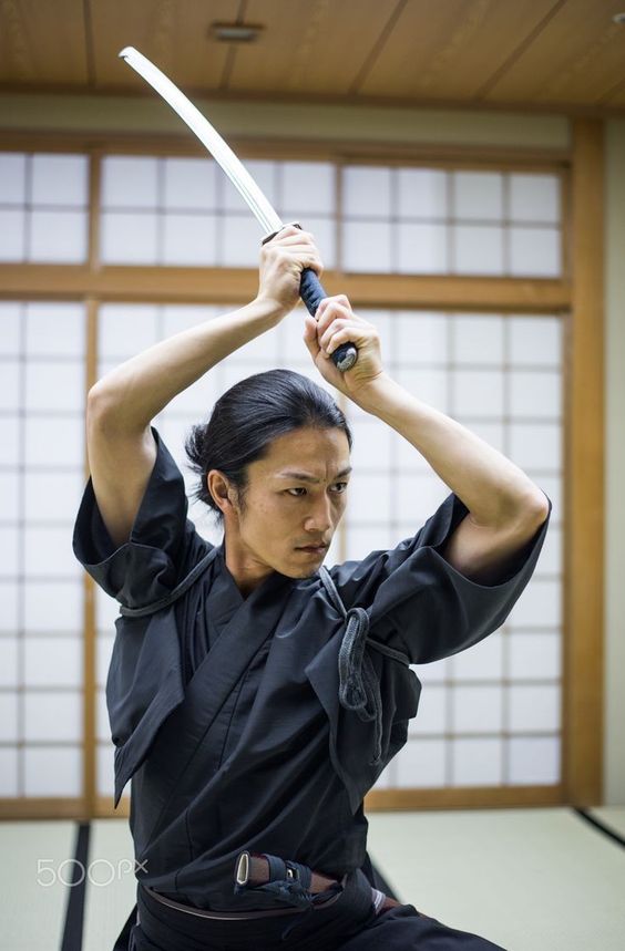 a young asian man in a traditional black martial arts uniform practices swordsmanship in a brightly lit dojo. he holds a katana above his head, poised to strike, his expression focused and determined. the image captures the discipline and grace of japanese martial arts against the simple backdrop of the training hall with its tatami mat floor and shoji screen doors.
