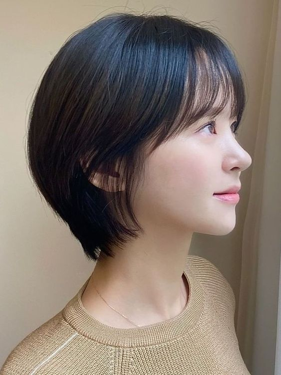 a young asian woman with short black hair, styled in a bob, is captured in a side profile portrait. the soft, natural lighting illuminates her face, highlighting her features. she is wearing a light brown sweater and her expression is neutral. the background is a plain wall, creating a minimalist aesthetic.