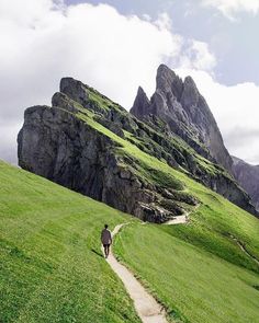 the image showcases a breathtaking mountain landscape with a lone hiker traversing a winding path. the path, carved into the lush green slopes, leads towards towering peaks that pierce the cloudy sky. the scene is bathed in natural daylight, illuminating the vibrant green of the grass and the rugged texture of the rocky mountainside.