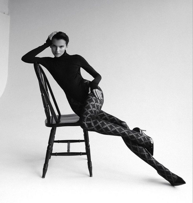 a black and white studio portrait of a woman. she is wearing a black turtleneck bodysuit, patterned tights, and high heels. she is sitting in a black chair, striking a pose with one hand on her head and her legs extended. the background is a neutral white.