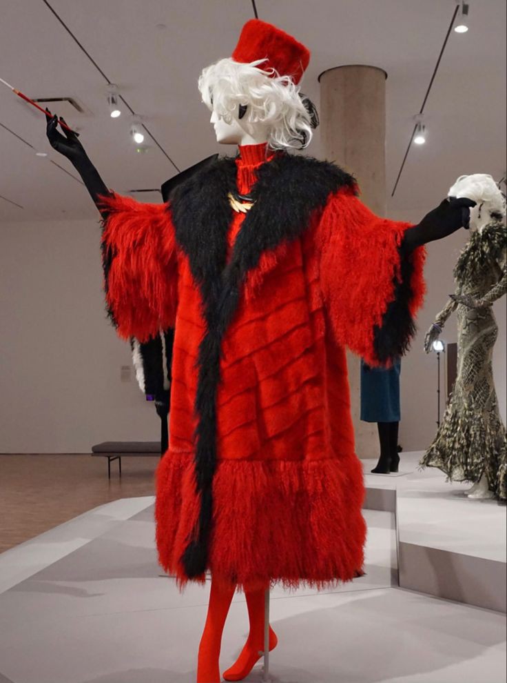 a fulllength shot of a mannequin dressed as cruella de vil from 101 dalmatians, standing in what appears to be a museum or exhibition hall. the mannequin wears a striking red fur coat with black trim, paired with matching red tights and heels. it has black gloves and holds a cigarette holder in its left hand. the mannequin sports a white wig and a red hat, completing the iconic villain look. other mannequins and displays are visible in the background.