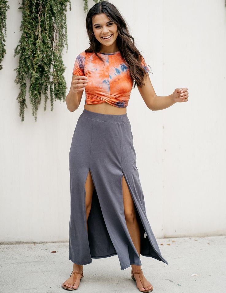 a young woman with long dark hair stands in front of a white wall, partially covered in greenery. she wears a vibrant orange and blue tiedye crop top and a long, dark gray maxi skirt with two thighhigh slits. she is smiling and appears happy and carefree. the bright, natural lighting enhances the summery vibe of the outfit.