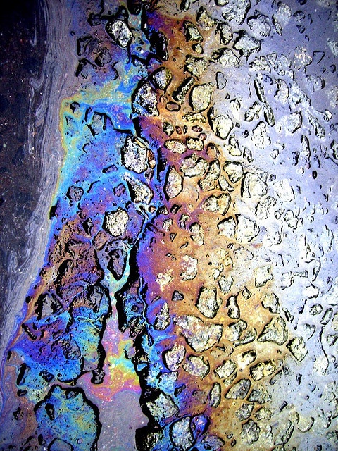 a closeup view of an oil slick on asphalt, showcasing the vibrant iridescent colors created by the thin film of oil. the intricate patterns and textures of the oil slick contrast with the rough surface of the asphalt, creating a captivating abstract composition. the image evokes a sense of both beauty and environmental impact.
