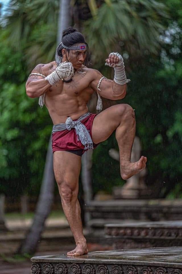 a young, muscular man wearing traditional muay thai shorts and a headband stands on a stone platform, preparing to strike. he has his right leg raised and is focused on his opponent, ready to unleash a powerful attack.  the man's intense expression and defined muscles convey his strength and determination. the lush green jungle setting adds an exotic and dramatic element to the scene.