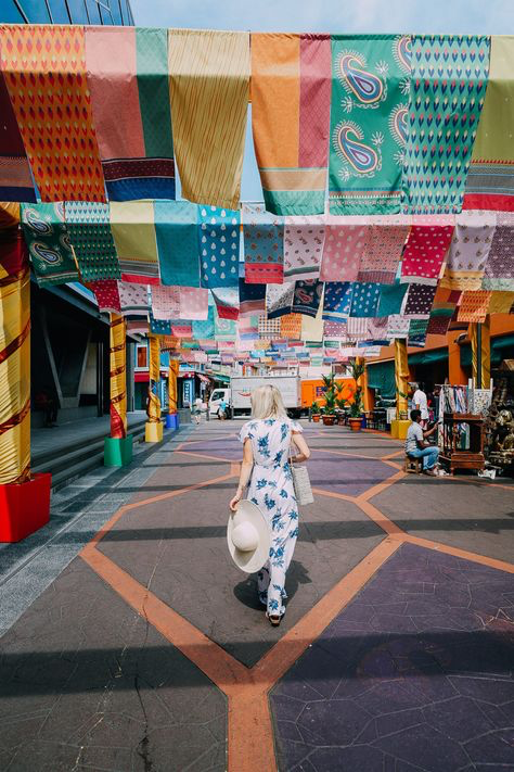 a woman in a floral dress walks down a colorful alleyway in little india, singapore. the alley is decorated with vibrant, patterned flags hanging overhead, creating a festive and cheerful atmosphere. shops and stalls line the sides of the street, and the overall scene is sunny and bustling with activity. the woman carries a large white sunhat.
