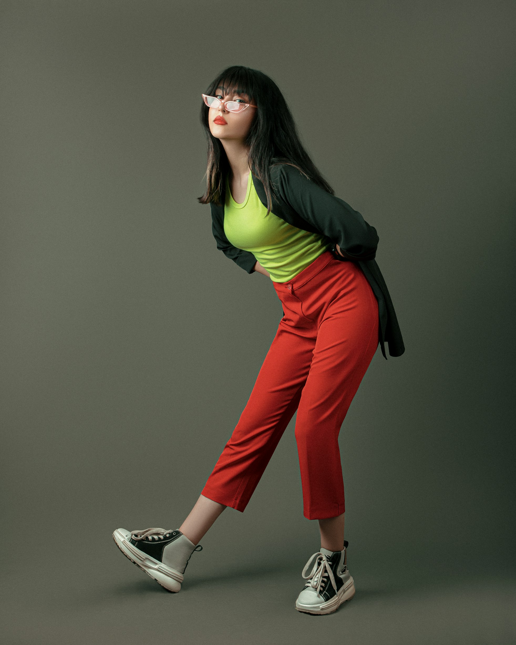 a young woman is posing in a studio. she has black hair with bangs and is wearing pinkframed glasses. she is wearing a neon green tank top, red capri pants, and a black blazer. her shoes are black and white platform high tops. she is standing with one leg extended and bent at the knee. she has her hands on her hips. the background is a solid gray.
