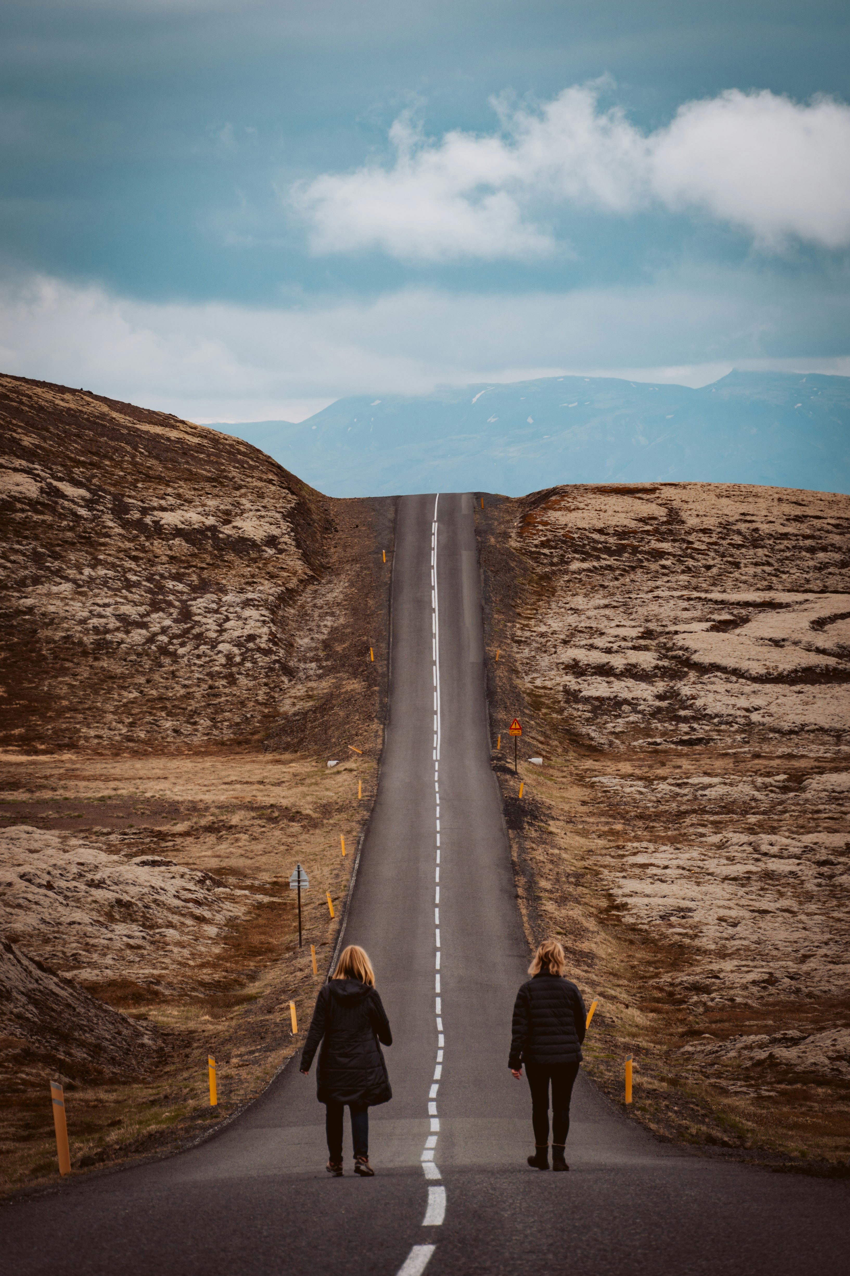 two women walk away from the camera down a seemingly endless straight road that cuts through a vast, mountainous landscape under a cloudy sky.  the women are wearing long coats and appear small against the grand scale of their surroundings.  the road is a stark contrast to the natural browns and greens of the hills, creating a strong leading line that draws the viewer's eye towards the distant mountains.