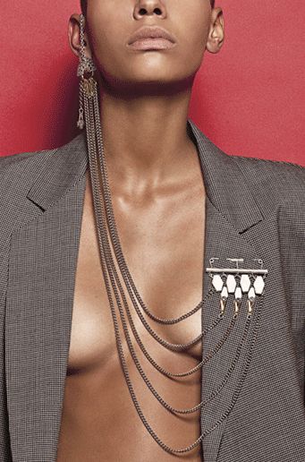 a closeup portrait of a woman with short hair wearing a gray blazer. she is barechested under the blazer. she is wearing a long, layered chain necklace that drapes down her chest and attaches to a silver and white brooch on the blazer. she is also wearing a long, dangling earring on her left ear. the background is a solid red color.