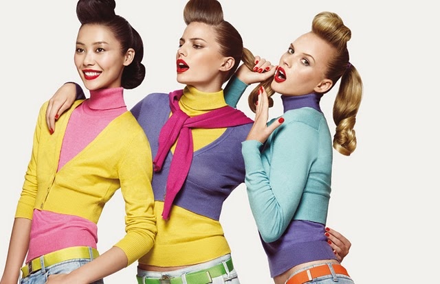 three women in retro 1950s style clothing are posing in a studio shot. they are wearing colorful layered sweaters and turtlenecks, with cropped cardigans. each woman has distinct hair and makeup, with vibrant red lipstick. they are wearing jeans with contrasting colored belts. the women display different poses, giving off a retro, fashionable vibe reminiscent of the 1950s and 1960s. one of the women is asian, while the other two appear to be caucasian.