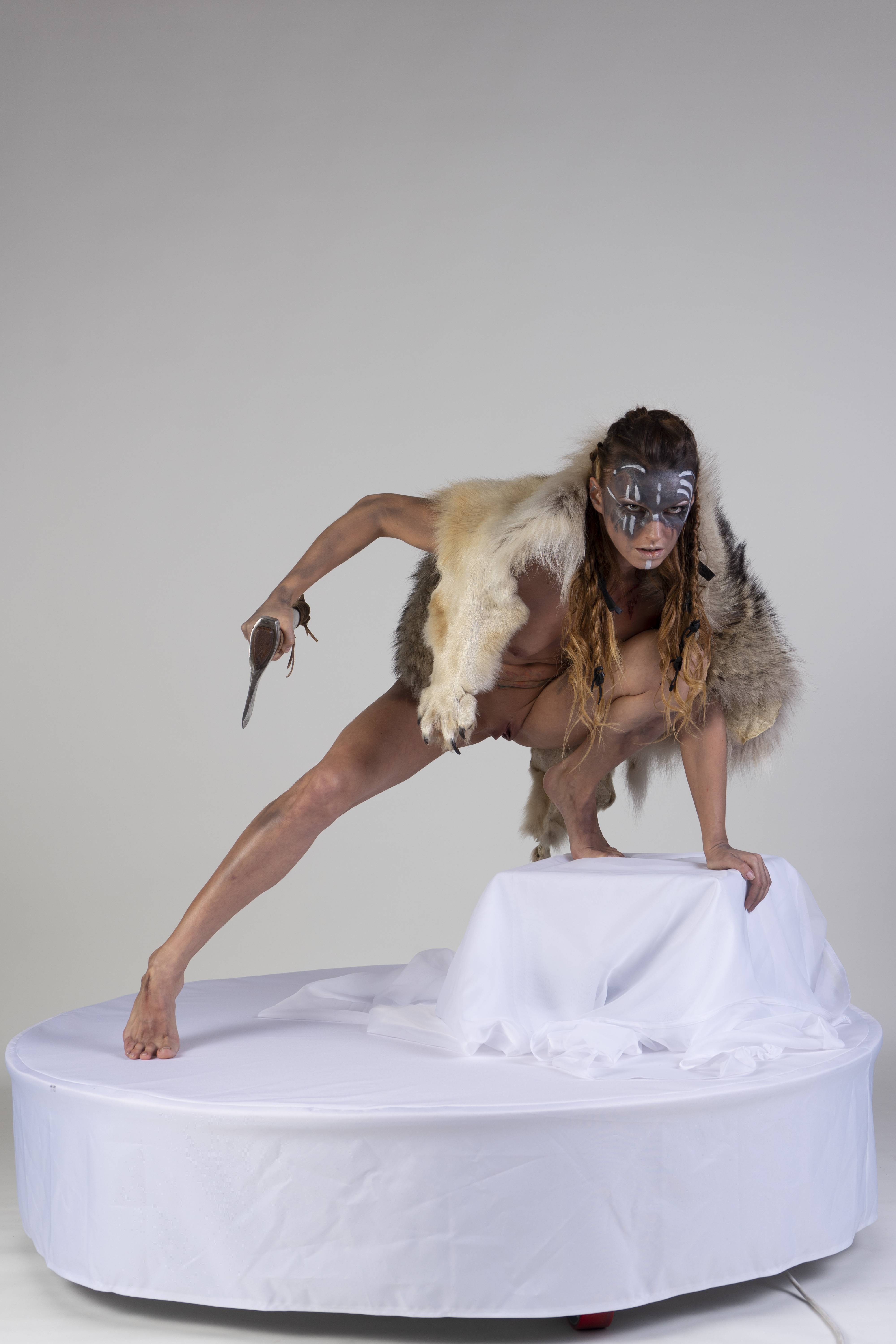 a woman dressed as a primitive tribal warrior is crouching on a white platform against a white background. she is wearing a fur loincloth and fur wrap, and has a mask painted on her face. she is barefoot and holding a knife. she is in a crouching pose, ready to attack.