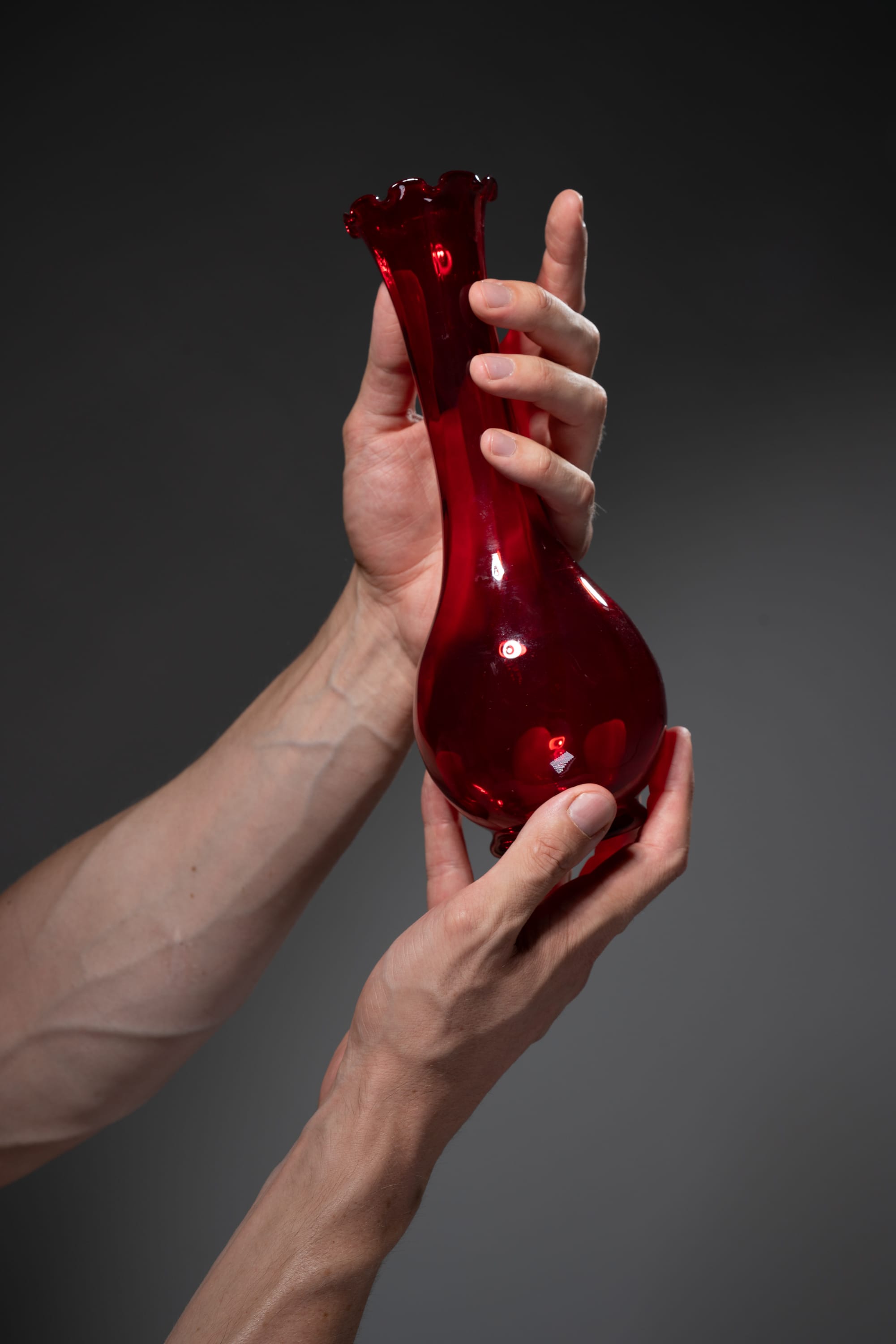 an adult is holding a red glass vase in his hands. the background is gray.