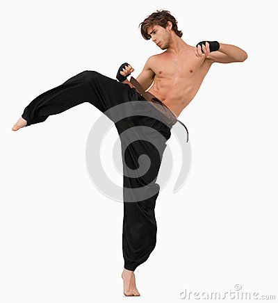 a muscular man wearing black martial arts pants and a black belt, with his torso bare, stands in a fighting pose, striking a leg out in front of him with his knee bent.  the man is midmotion, his arms are out. the background is white.