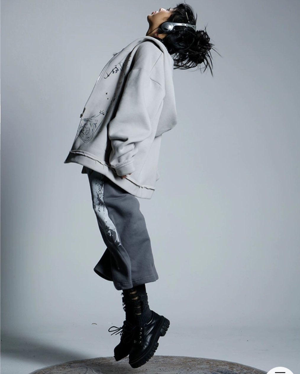 the image shows a stylish young woman, possibly a fashion model, midjump.  she is wearing an oversized light grey hoodie with subtle graphic prints, paired with dark grey wideleg sweatpants and chunky black combat boots. she's wearing headphones around her neck and her hair is styled in a loose updo. the background is a simple, light grey studio backdrop. the overall impression is one of casual, yet fashionable streetwear style. the pose and lighting suggest a fashion shoot or editorial style photograph.