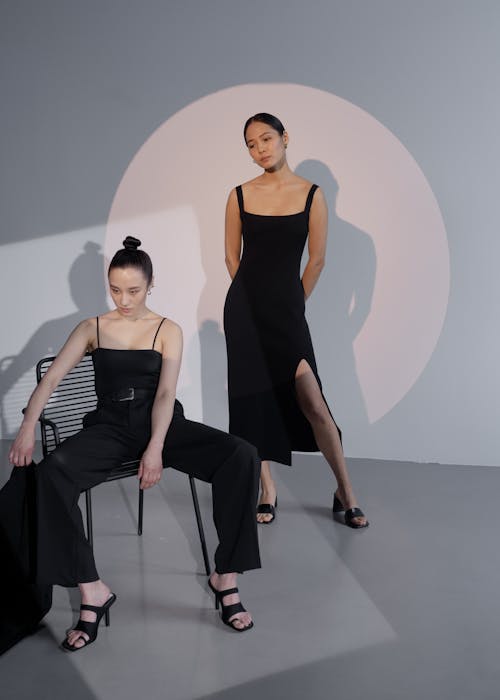 two women models are showcased in a minimalist studio setting. one model is seated on a black metal chair wearing a black pantsuit and black heeled sandals.  the other model stands behind her wearing a black midi dress with a thighhigh slit, also wearing black heeled sandals. both outfits are simple, elegant, and contemporary in style. a large, soft, circular light source enhances the overall aesthetic. the image is clean and sharply focused, highlighting the clothing and the models' poses. the background is a muted gray, and the overall atmosphere is calm and sophisticated.
