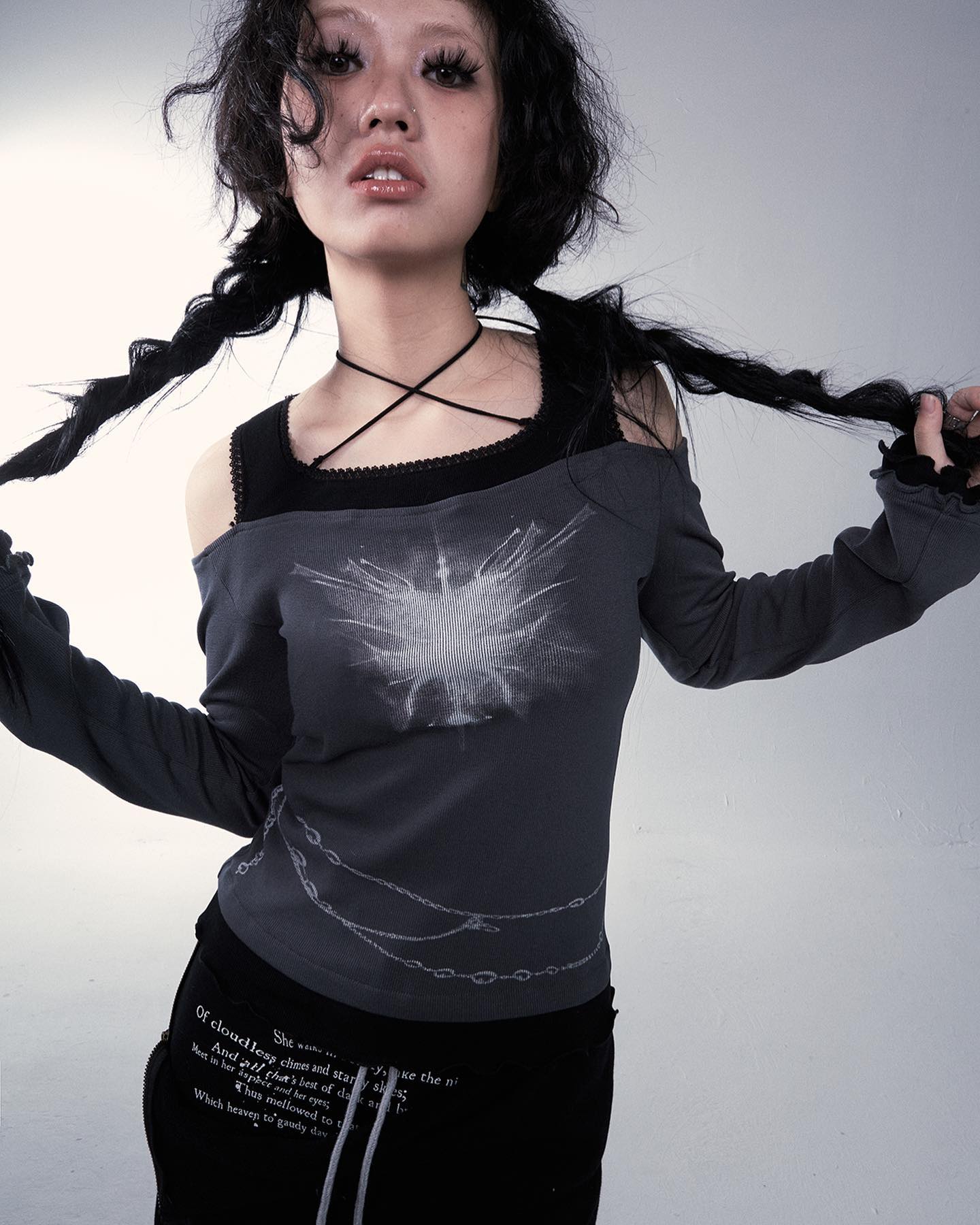 the image is a studio shot of a young woman modeling dark fashion attire. she has dark hair styled in long braided pigtails, pale skin and is wearing an offshoulder gray longsleeved top with a graphic of chains and a white, abstract design. the top is paired with black pants that also have a subtle textual print. she's also wearing a black choker necklace. the overall style is dark academia meets goth/grunge with an edgy and alternative feel. the background is plain and light gray, allowing the clothing and model to stand out prominently.