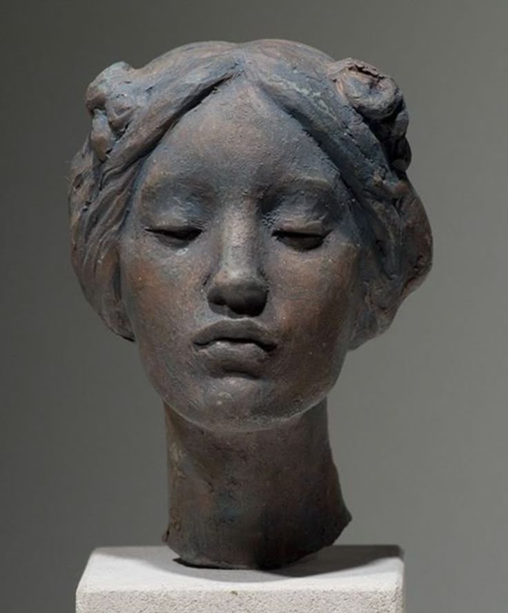 the image is a closeup studio shot of a sculpture bust of a woman's head. the sculpture is made from a dark, earthy material, possibly clay or stoneware, and exhibits a classical aesthetic. the woman has her eyes closed, conveying a sense of serenity and peace. her facial features are subtly detailed, showcasing a delicate and artistic rendering. the sculpture is displayed indoors on a lightcolored stone stand, enhancing the artwork's presentation.