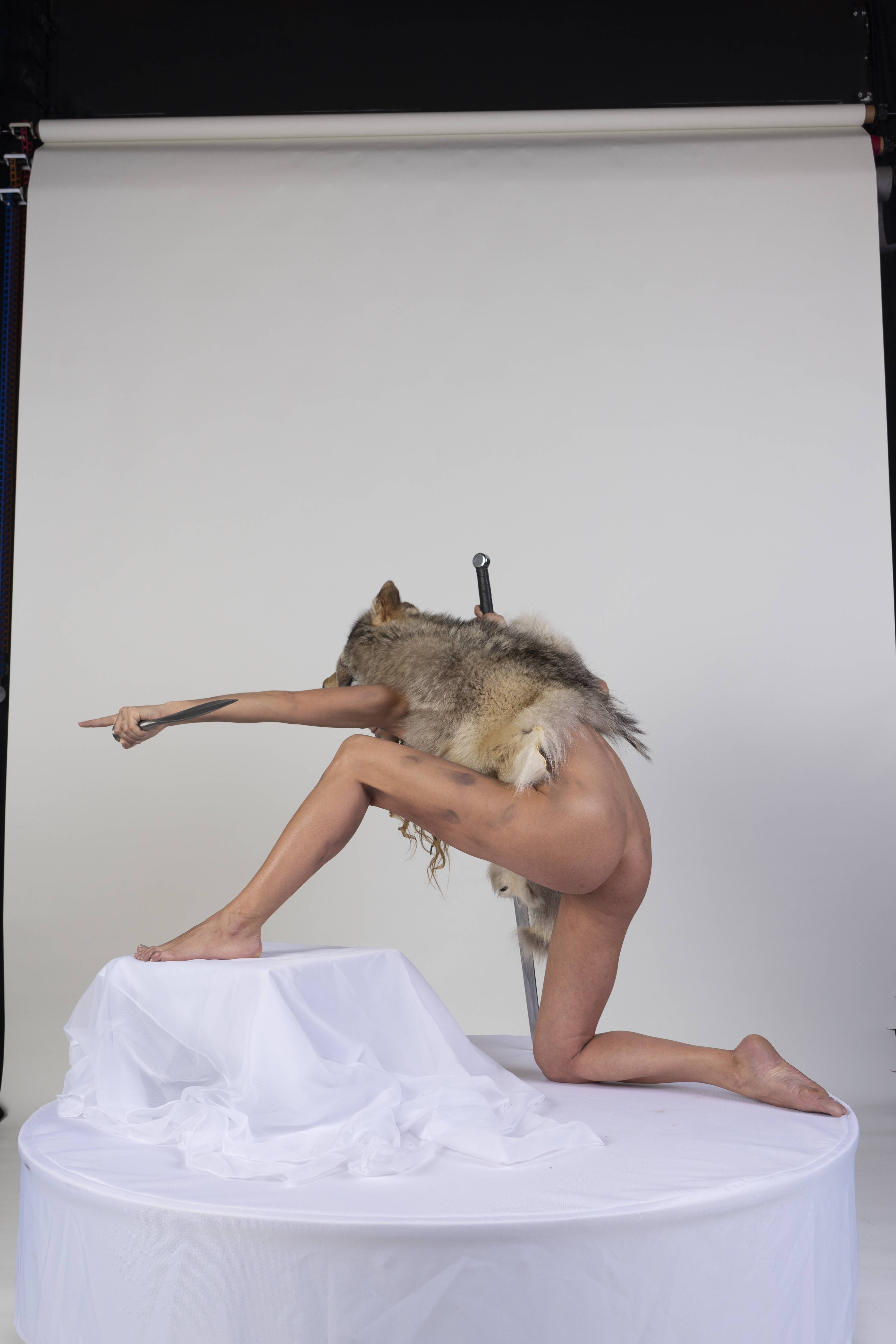 a nude woman poses on a white platform. she is draped with fur from a wolf and has a sword behind her. she points with a knife in her hand.