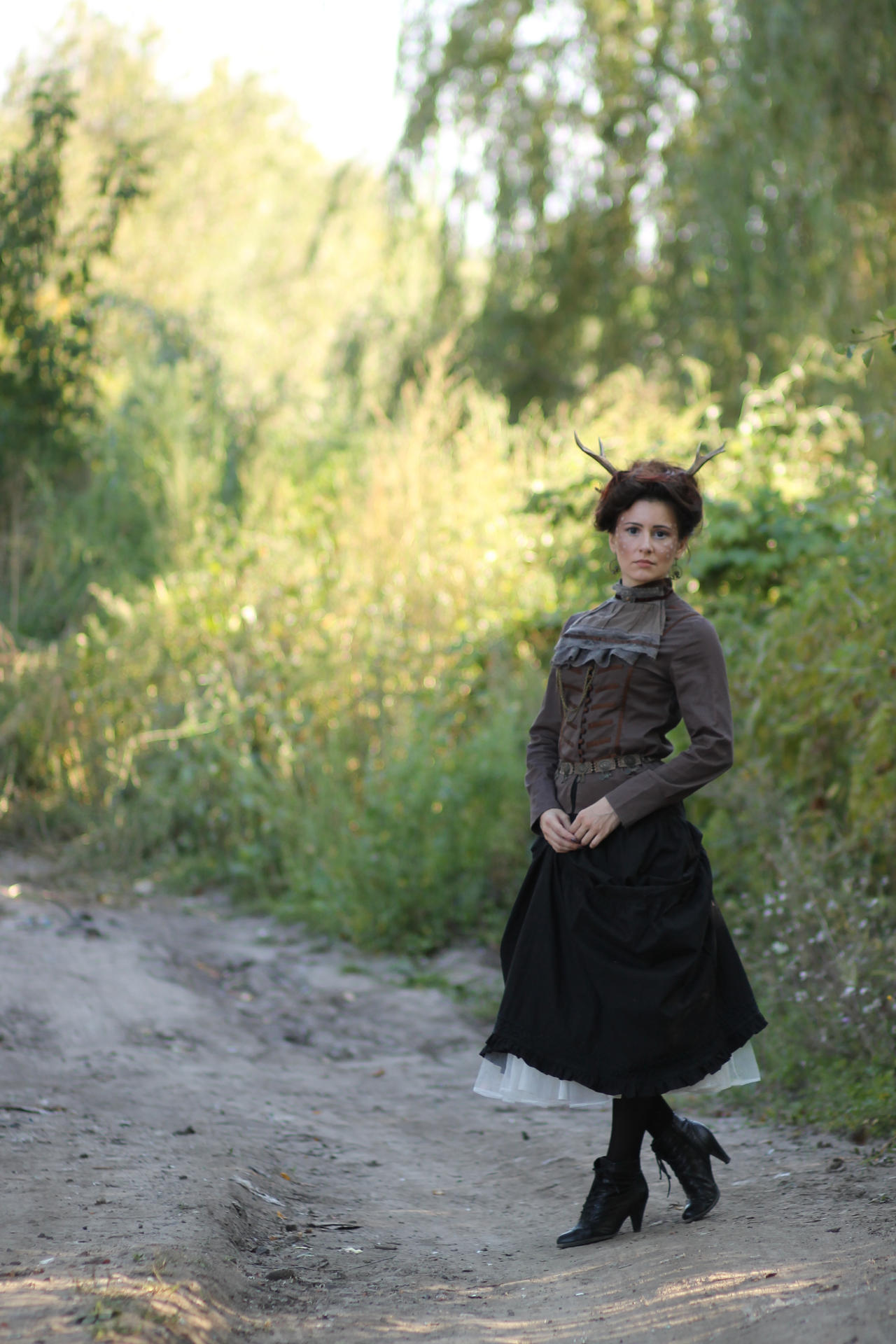 a fulllength portrait of a woman in a vintageinspired steampunk costume. she's wearing a brown jacket and a long black skirt with a white petticoat underneath.  antlers are adorn her hair as a headpiece. she is standing on a dirt path in a forest, surrounded by lush greenery. the overall style is evocative of a whimsical, fantastical scene.