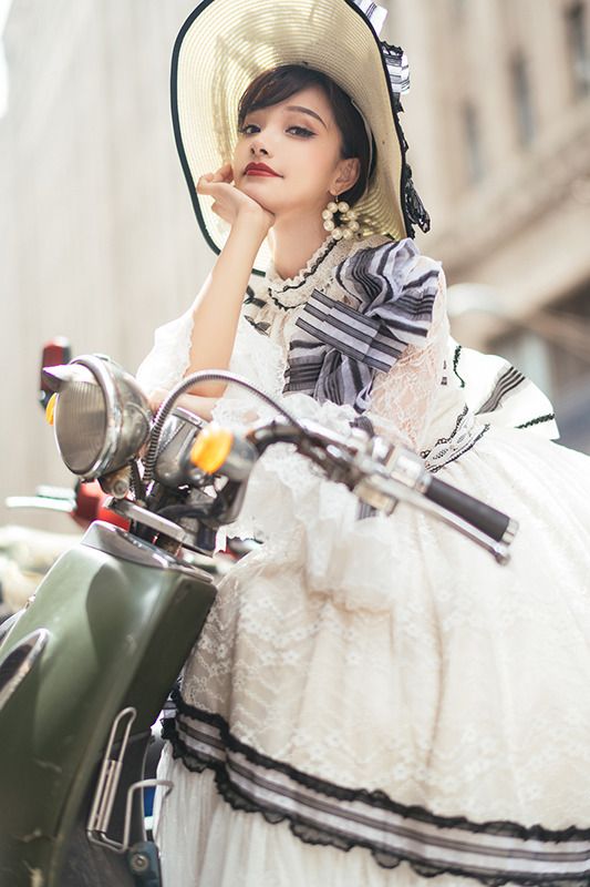 a woman wearing a white dress and a straw hat sits on a vintage scooter in a city street. she has her hand on her chin and is looking at the camera. the image is shot in a vintage style.