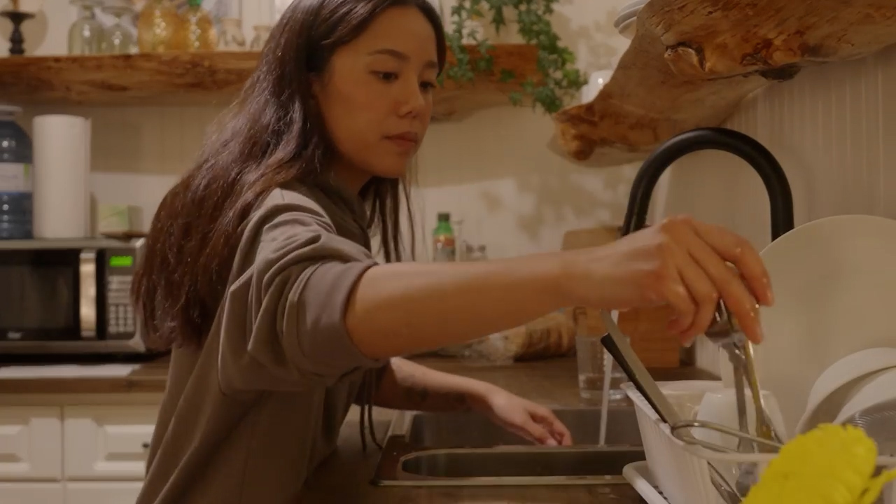 a young woman with long brown hair is washing dishes in a kitchen sink. she is wearing a brown sweatshirt and is standing at the sink, which is filled with water and dishes. the kitchen is welllit and has a warm and inviting atmosphere, featuring white cabinets and wooden countertops.